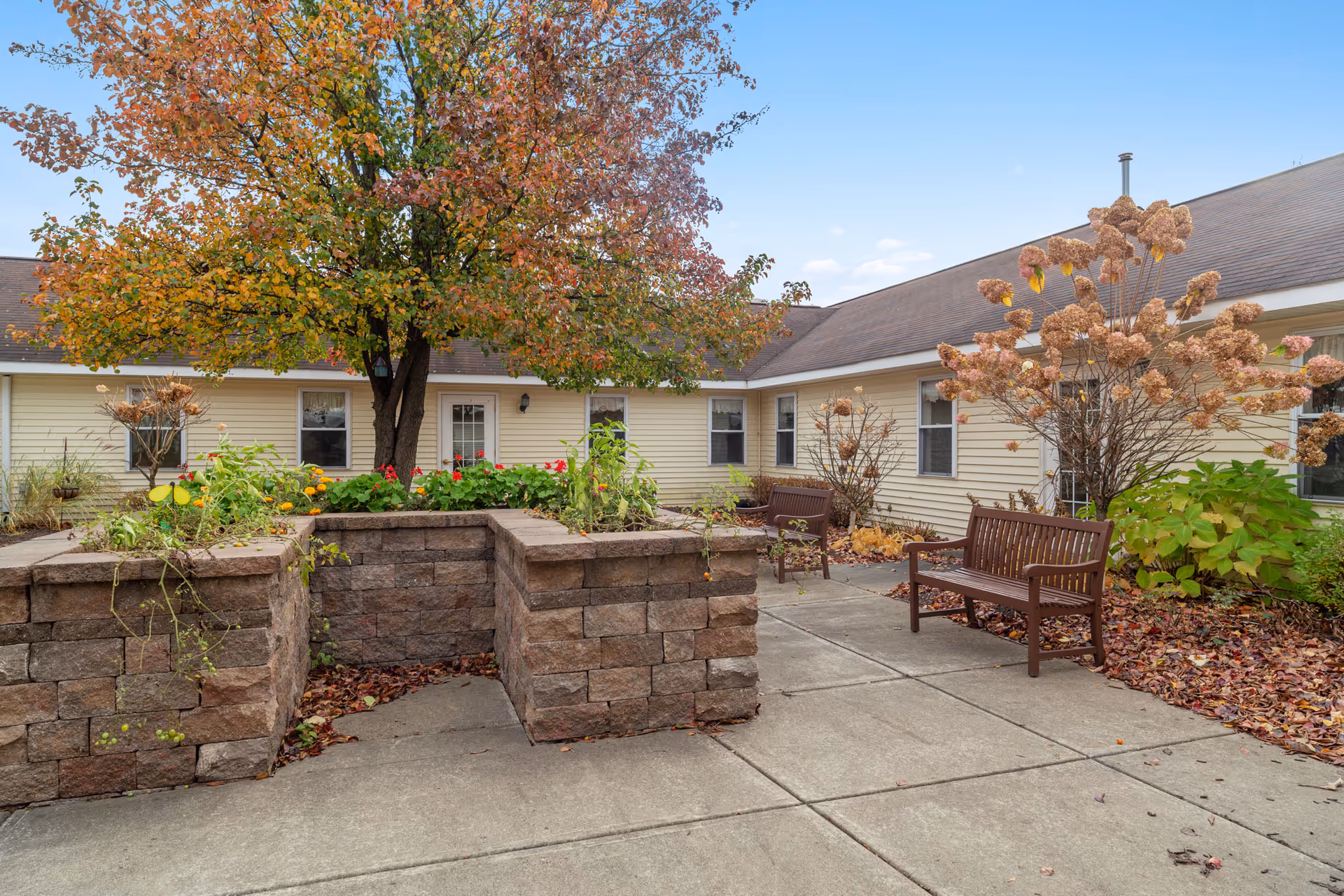 Courtyard patio with stone planters, wooden benches, autumn trees and a surrounding single-story building.