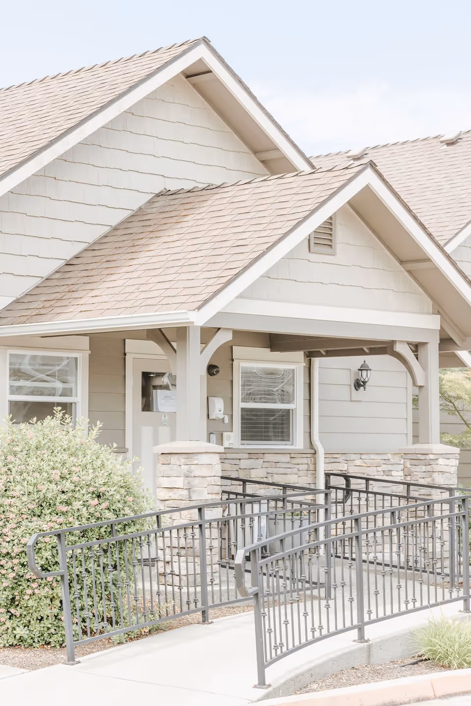 Exterior view of a light-colored building with a covered entrance, stone pillars, and a black metal wheelchair ramp leading to the door. There are two windows and a wall-mounted light fixture near the entrance, with some greenery and bushes nearby.