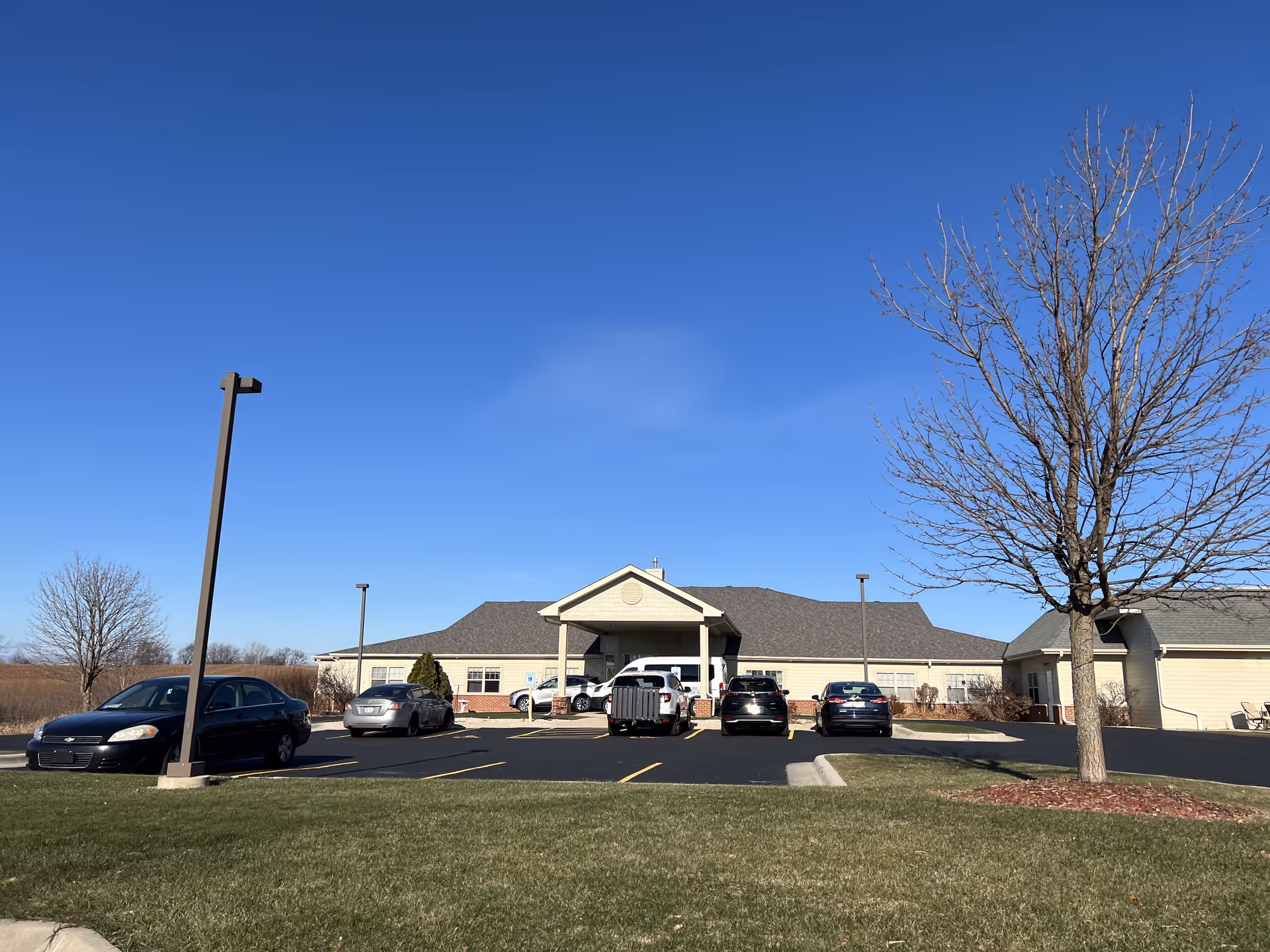 Exterior view of a single-story building with a covered entrance and several parked cars in front. The sky is clear and blue, and there are leafless trees and a grassy area surrounding the parking lot.
