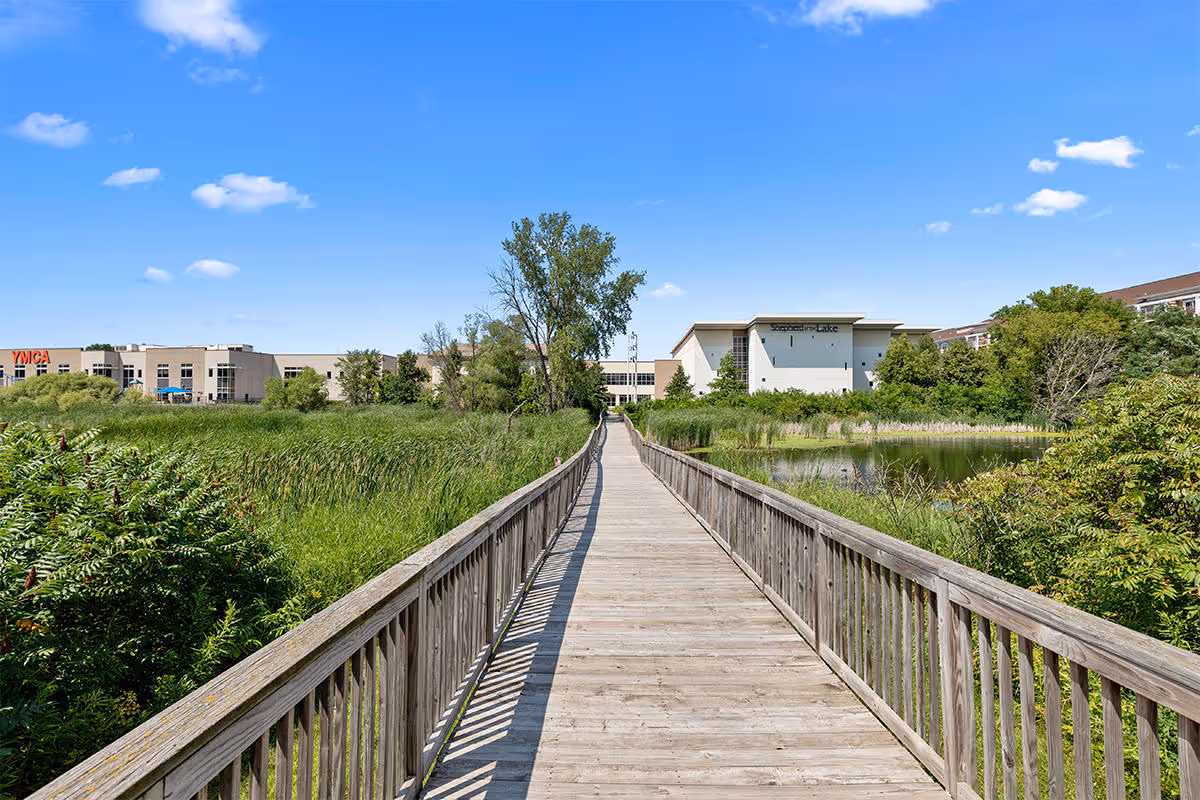 A wooden boardwalk stretches forward through lush green vegetation and wetlands under a bright blue sky with scattered clouds. In the distance, there are buildings including one with a YMCA sign and another labeled 'Sugarcreek Lake'.