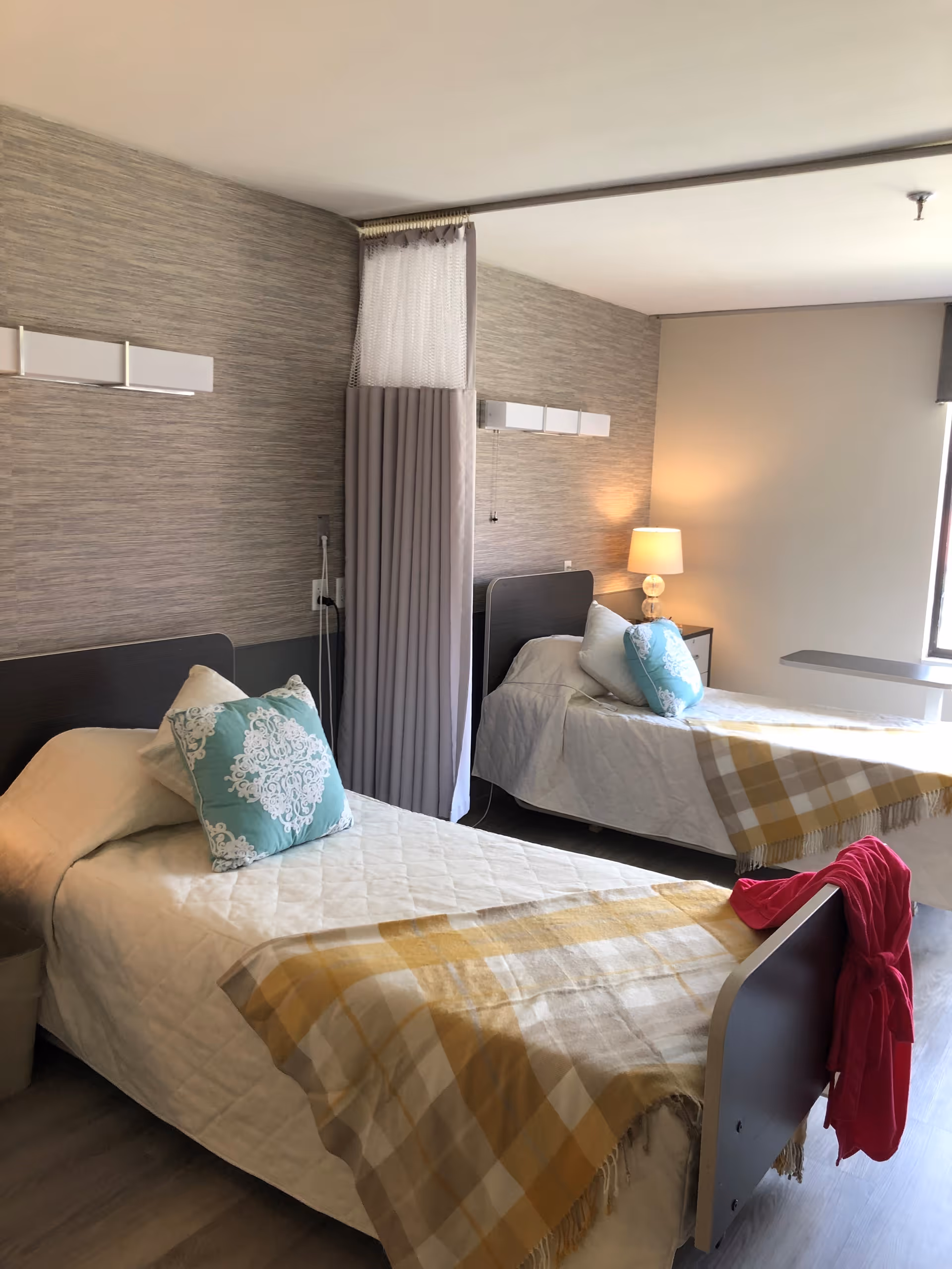 A bright room with two single beds, each with a white quilted bedspread, a decorative blue pillow, and a yellow plaid throw blanket. A bedside table with a lamp is positioned between the beds, and a privacy curtain hangs from the ceiling. A red garment is draped over the footboard of one bed. The room has light-colored walls and a window letting in natural light.