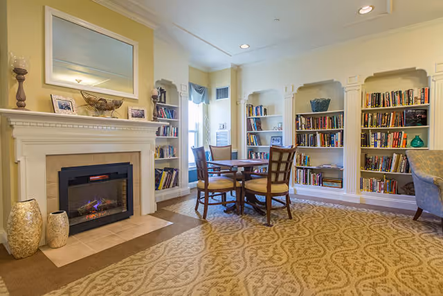 A cozy room with a fireplace, a round wooden table with four chairs, built-in bookshelves filled with books, and a patterned carpet. The walls are painted light yellow, and there is a large mirror above the fireplace.