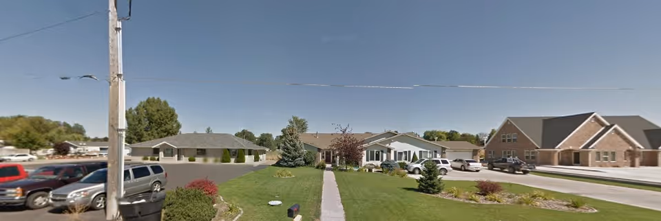 Front exterior view of single-story assisted living buildings with a central sidewalk, landscaped lawn and parked cars.