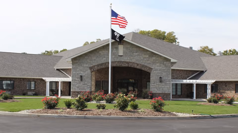 Front exterior view of Cedarhurst of Breese facility with a stone and brick facade, a covered entrance, two flagpoles with the American flag and another flag, surrounded by green lawn and flower beds.