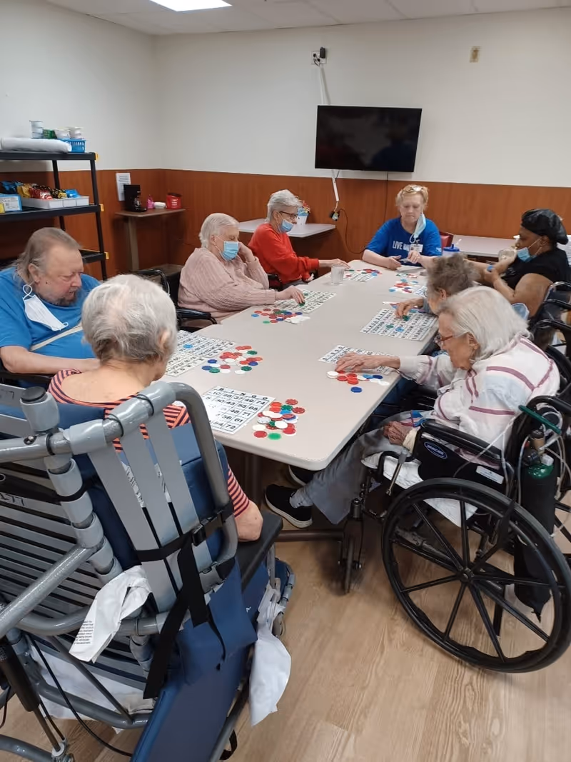 A group of elderly individuals sitting around a rectangular table playing bingo. Some are in wheelchairs and others are seated in chairs. They are in a room with wood-paneled walls and a television mounted on the wall. Several people are wearing face masks.
