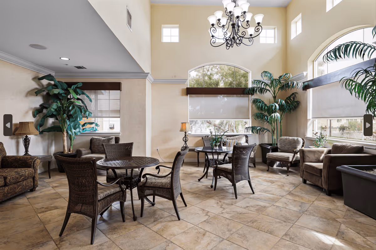 A bright and spacious common area in an assisted living facility with high ceilings and large windows. The room features several round tables with wicker chairs, comfortable armchairs, potted plants, and decorative lamps. The floor is tiled, and natural light fills the space through the windows.