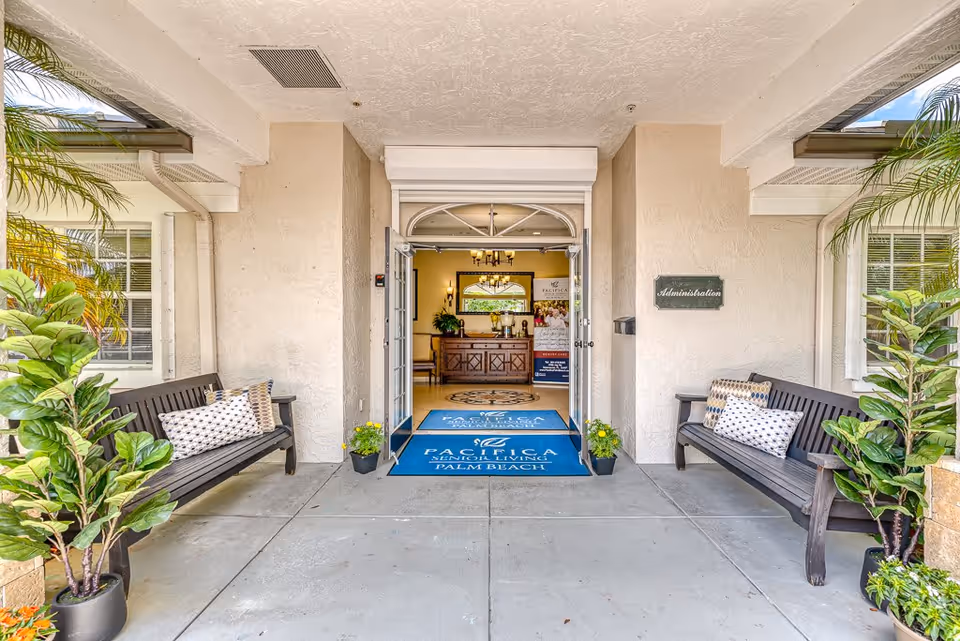 Entrance to a senior living facility with two wooden benches on either side, each with decorative pillows. There are potted plants near the benches and a blue mat at the entrance that reads 'Pacifica Senior Living Palm Beach'. The double glass doors are open, revealing a reception area inside with a wooden desk, plants, and a banner.