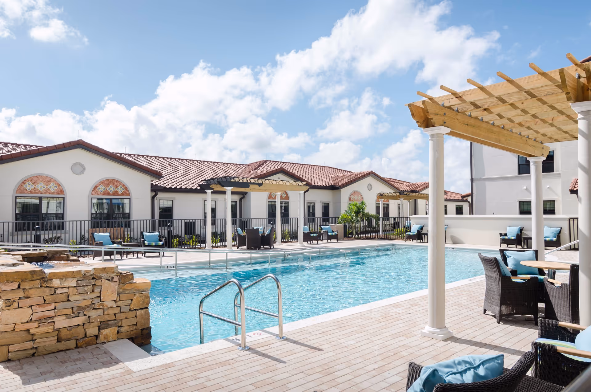 Outdoor swimming pool area at American House Coconut Point with clear blue water, surrounded by a tiled deck with black wicker chairs and tables with blue cushions. There are pergolas with wooden beams and white columns providing shade. The background shows a building with white walls, red-tiled roofs, and decorative arched windows under a partly cloudy blue sky.