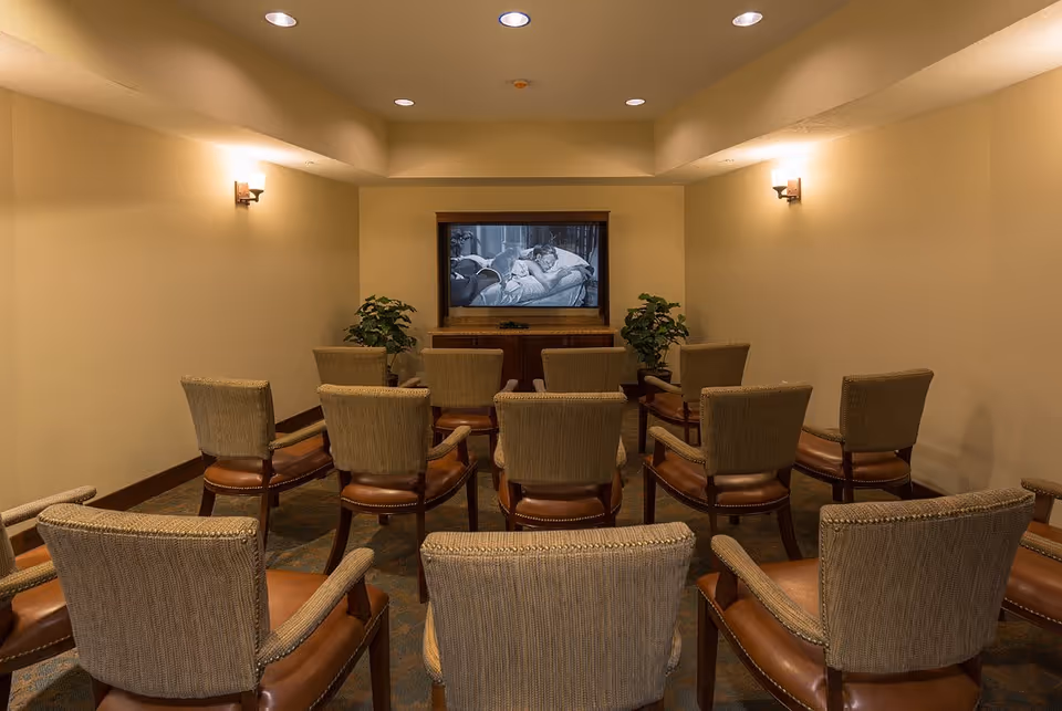 Small theater room with several rows of upholstered chairs facing a large screen showing a black and white movie. The room has beige walls, wall sconces for lighting, and two potted plants on either side of the screen.