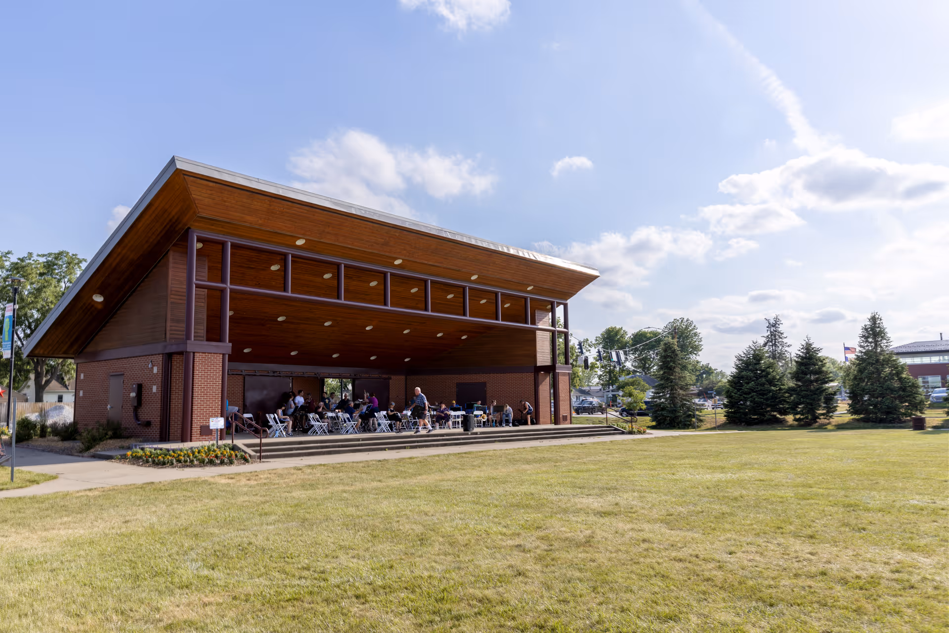 Large open-air wooden pavilion with people seated inside facing a grassy lawn under a blue sky.