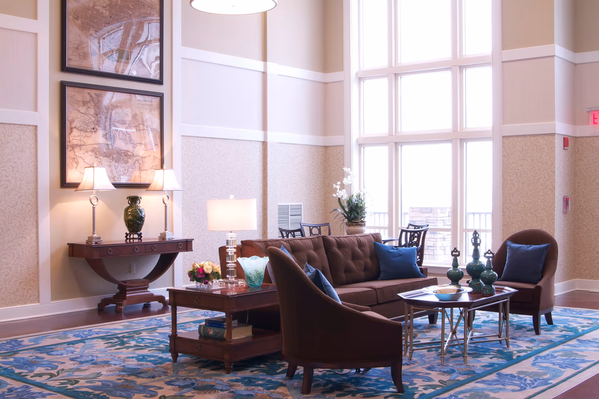 A bright and elegant living room area with a large window letting in natural light. The room features a brown tufted sofa with blue pillows, two matching armchairs, a wooden coffee table with decorative green vases, and a side table with a lamp and flowers. There is a wooden console table against the wall with two lamps and a green vase, and two framed maps hanging above it. The floor is covered with a blue patterned rug.