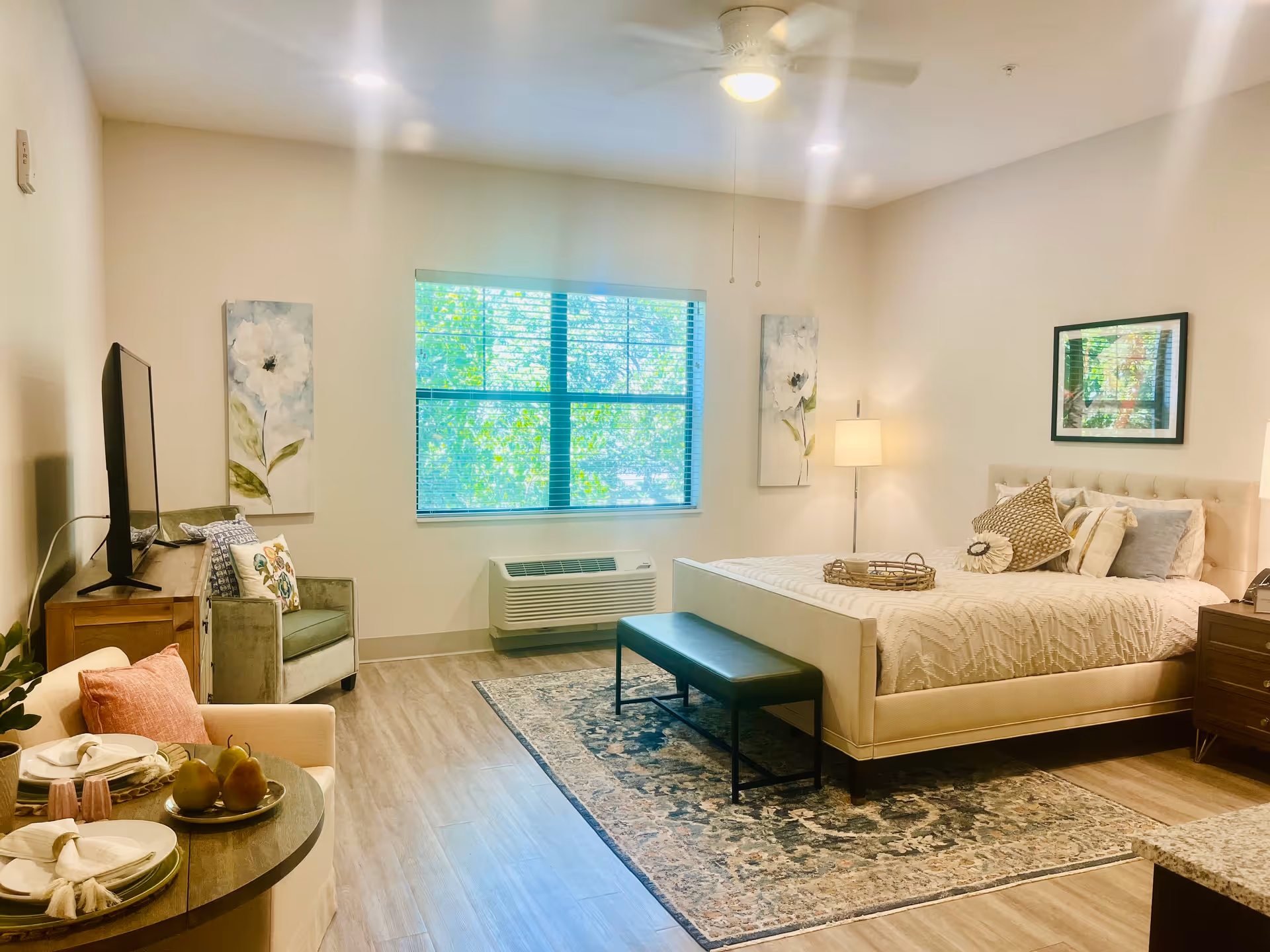 A bright and spacious bedroom with a large window letting in natural light. The room features a comfortable bed with multiple pillows, a bench at the foot of the bed, a patterned area rug, a floor lamp, and framed artwork on the walls. There is also a TV on a wooden stand, an armchair with decorative pillows, and a small round table set with plates and pears. The flooring is wood-style, and the walls are painted light beige.