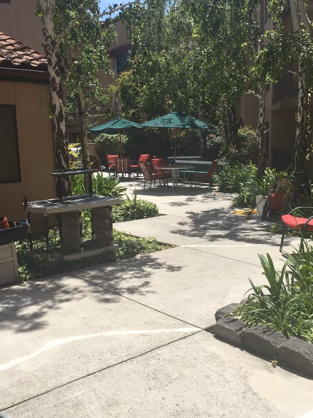 Outdoor patio area with concrete walkways, green umbrellas shading tables and chairs with red cushions, surrounded by trees and plants in a garden setting.