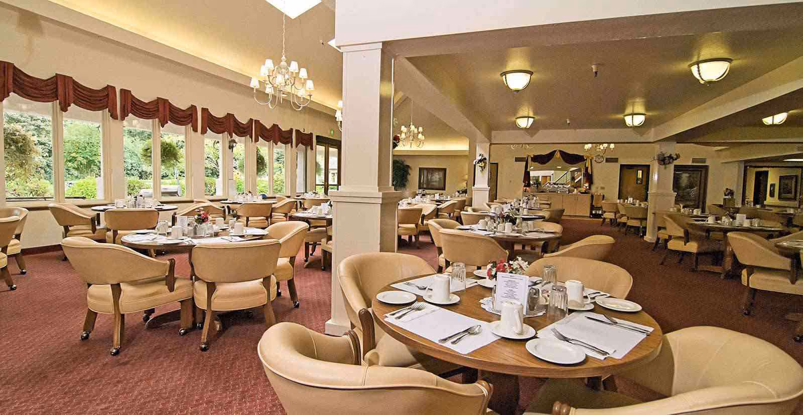 A spacious dining room with multiple round tables set with white plates, cups, silverware, and napkins. The room features beige upholstered chairs, large windows with red valances letting in natural light, chandeliers, and a carpeted floor. There are decorative plants and a buffet area in the background.