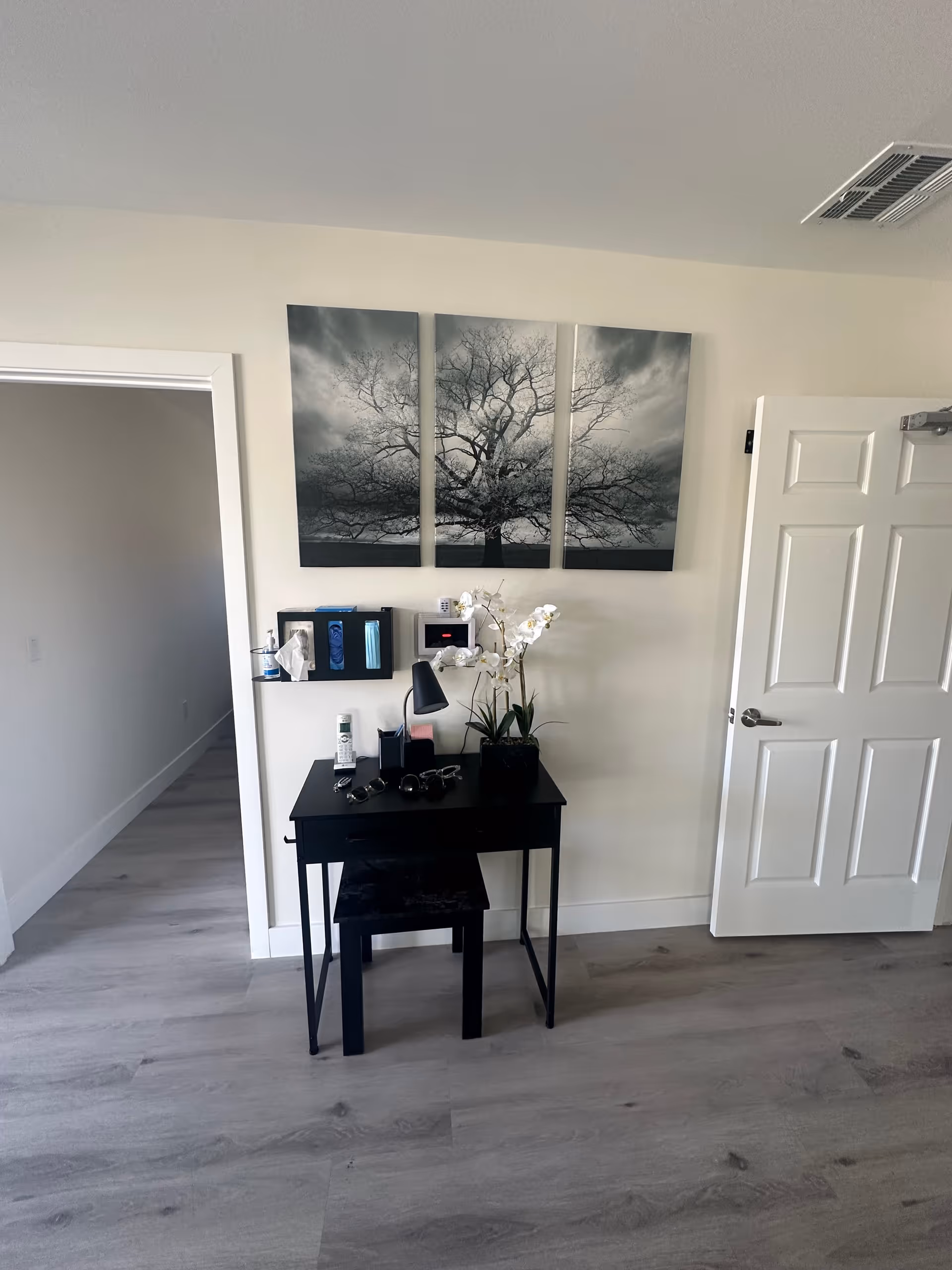 A small black desk with a matching stool is placed against a white wall. On the desk are a black lamp, a potted white orchid, a pair of sunglasses, a bottle of hand sanitizer, and some other small items. Above the desk hangs a three-panel black and white artwork of a large tree with bare branches. To the right is a white door slightly ajar, and to the left is an open doorway leading to a hallway. The floor is light-colored wood.