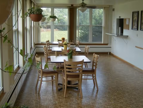 A bright dining area with several wooden tables and chairs arranged neatly. There are potted plants on the tables and hanging plants near large windows that let in natural light and offer a view of greenery outside. The floor has a patterned design, and the walls are light-colored with a bulletin board and framed pictures.