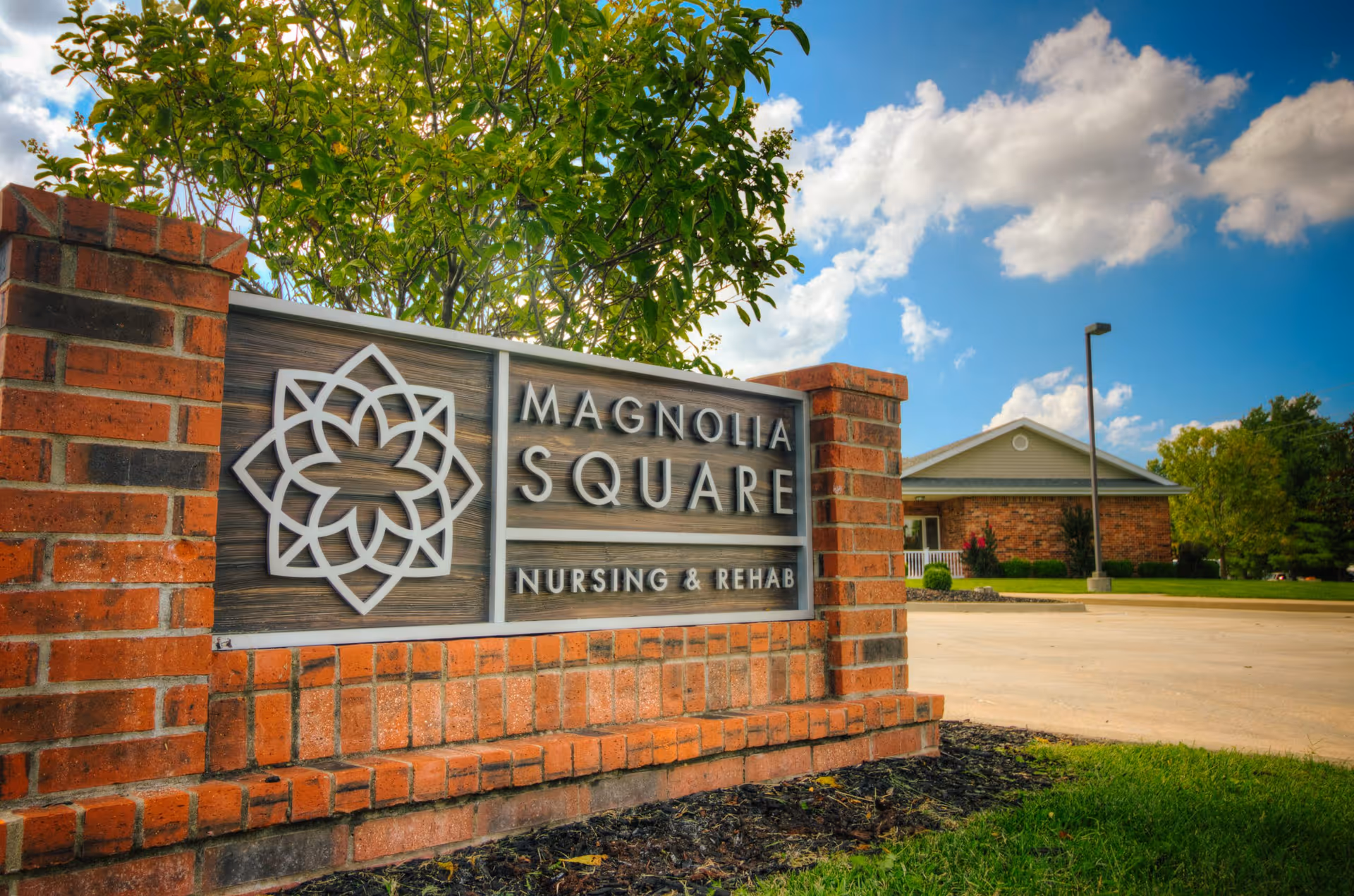 Brick sign for Magnolia Square Nursing & Rehab with a decorative floral logo, set outdoors with a building, trees, and a partly cloudy sky in the background.