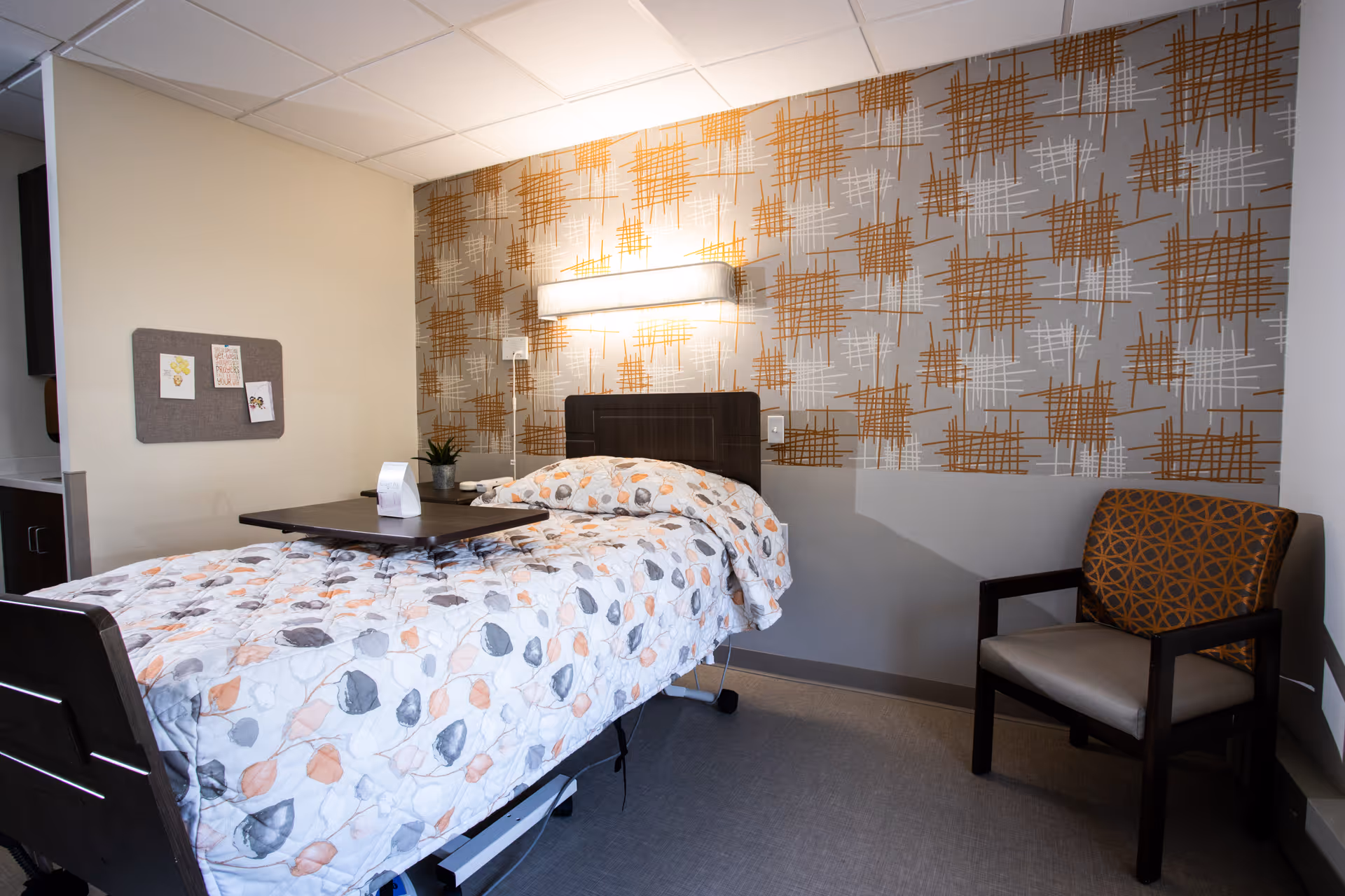 A single bed with a floral patterned comforter in a room with a patterned accent wall. There is a dark wooden headboard, a wall-mounted light above the bed, a small plant on a bedside table, a chair with an orange and brown geometric pattern cushion, and a small bulletin board with pinned notes on the wall.