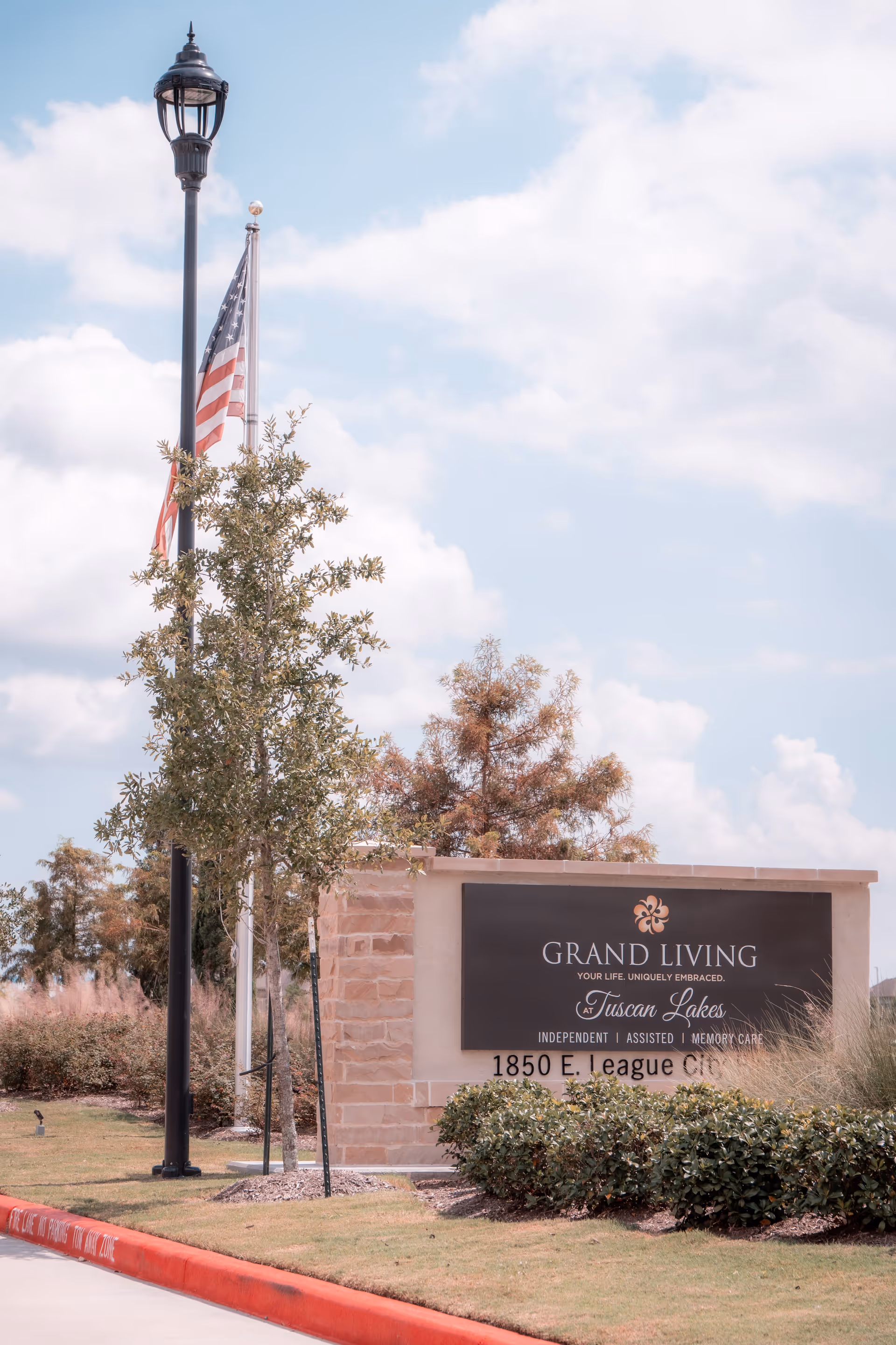 Outdoor view of the entrance sign for Grand Living at Tuscan Lakes, featuring a stone and stucco sign with the facility's name and address, surrounded by landscaping, a street lamp, and an American flag against a partly cloudy sky.