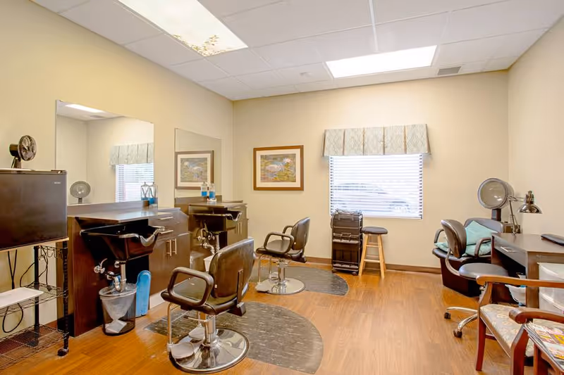 Interior of a hair salon area in an assisted living facility with two salon chairs in front of mirrors, a small refrigerator, a window with blinds and a valance, framed artwork on the wall, and various salon equipment including hair dryers and a rolling cart.