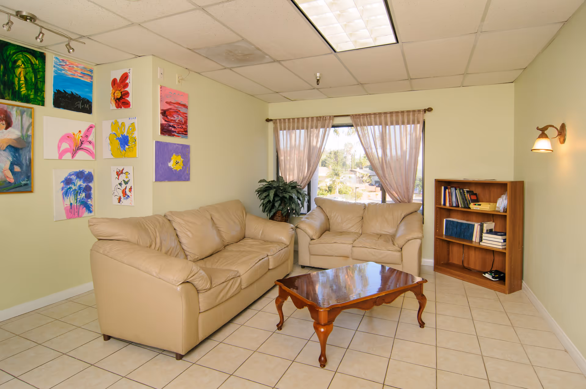 A cozy living room with two beige leather sofas arranged around a wooden coffee table. The room has light green walls adorned with colorful paintings on one side. A window with sheer curtains lets in natural light, and there is a small wooden bookshelf with books and a lamp on the right side. The floor is tiled, and a wall-mounted light fixture is visible on the right wall.