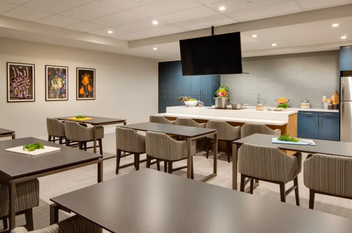 A modern dining area with several dark rectangular tables and striped upholstered chairs. In the background, there is a kitchen area with blue cabinets, a white countertop island with bar stools, a large flat-screen TV mounted above the island, and various kitchen items including pots, bowls, and a fruit basket. Three framed abstract art pieces hang on the wall to the left.