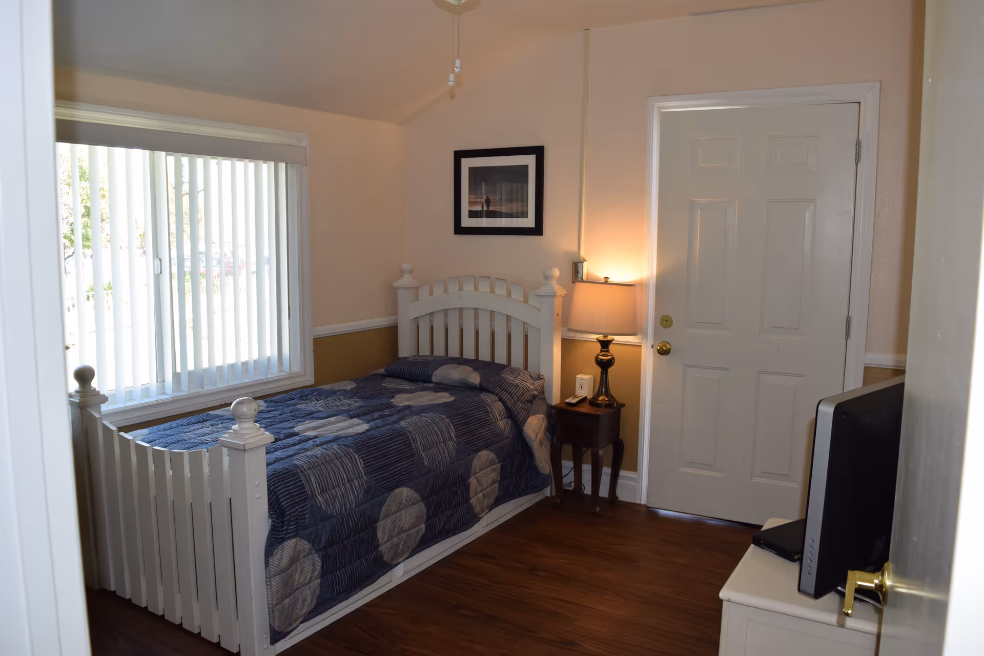 A small bedroom with a white wooden bed frame and a blue patterned comforter. There is a window with vertical blinds on the left wall, a framed picture above the bed, a wooden nightstand with a lamp next to the bed, and a closed white door on the right wall. A television is placed on a white dresser in the foreground on the right side.
