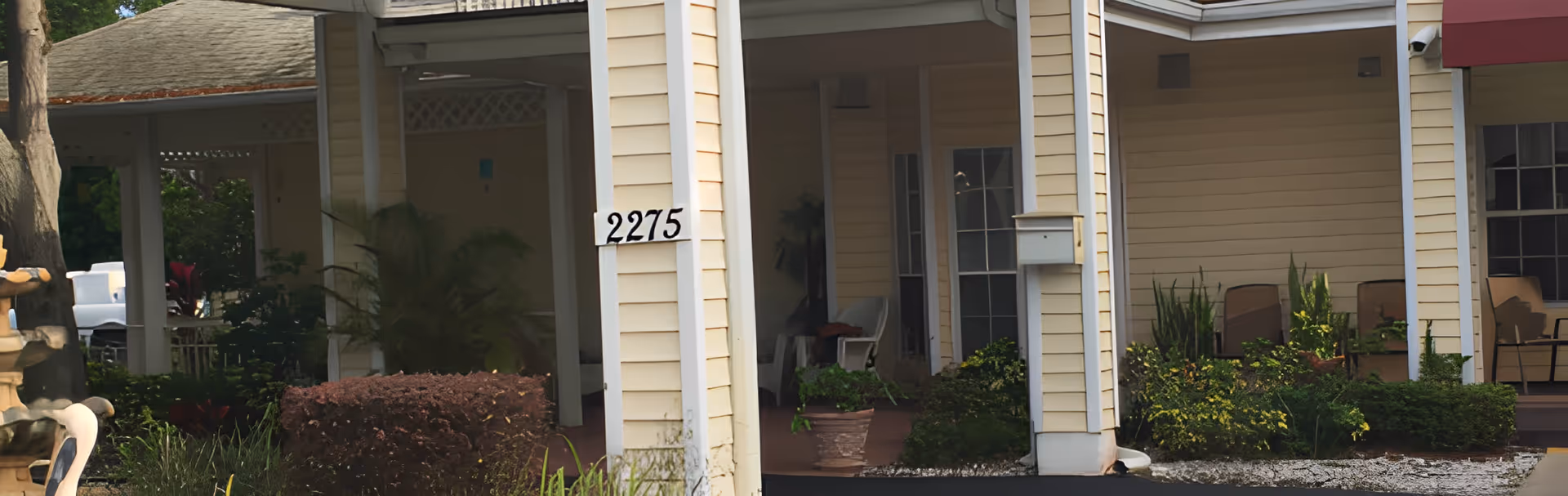 Exterior view of a building with beige siding and white columns, featuring the address number 2275. There are plants and shrubs in front of the building, and a covered porch area with chairs is visible.
