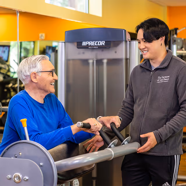 An elderly man in a blue long-sleeve shirt using a Precor multi-press exercise machine while smiling and interacting with a younger male staff member wearing a gray jacket with The Terraces of Los Gatos logo in a gym setting with yellow walls and mirrors.