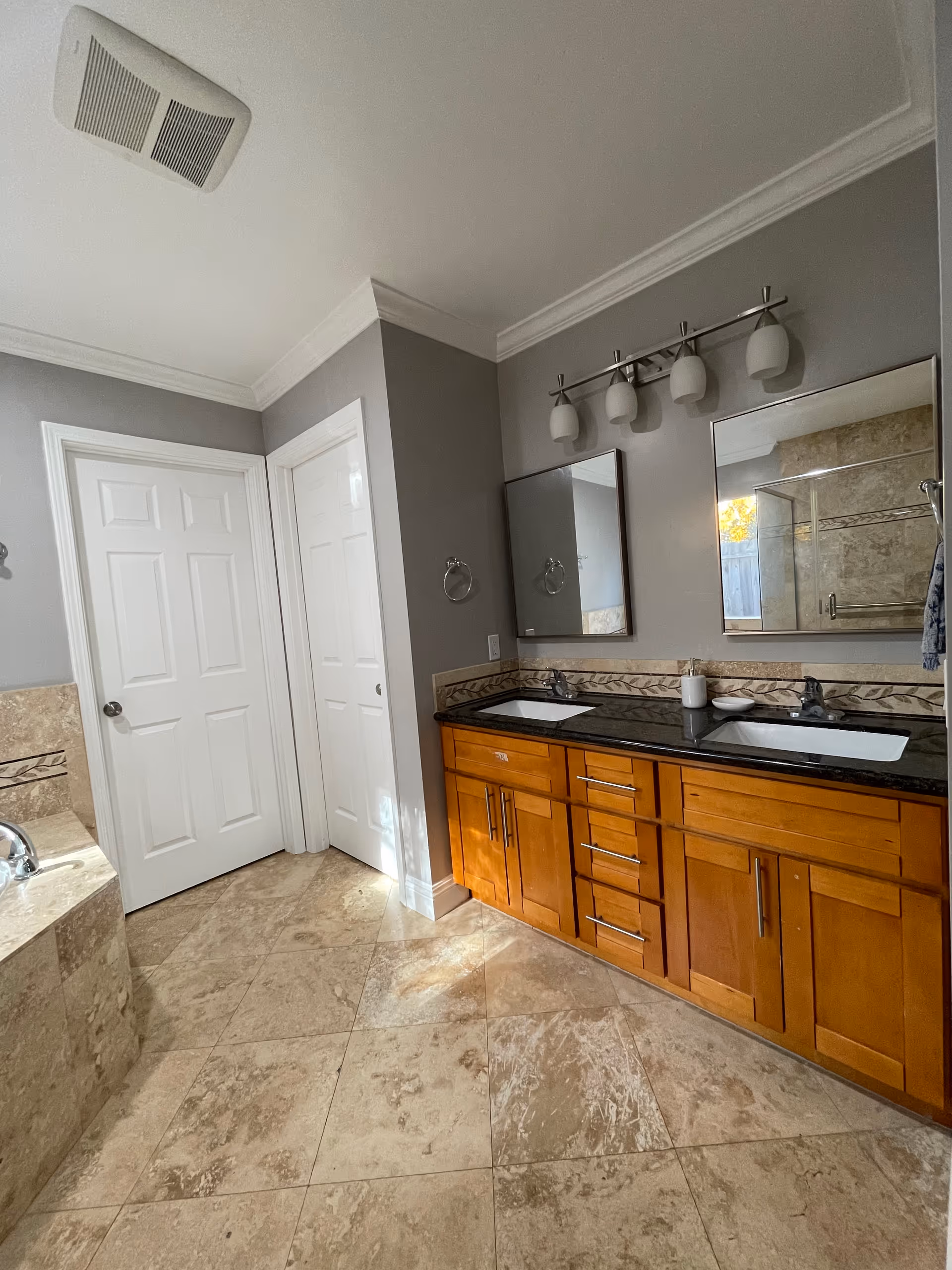A bathroom with beige tiled floor and walls, featuring a double sink vanity with wooden cabinets and black countertop. Above the sinks are two mirrors and a light fixture with four frosted glass shades. To the left, there is a built-in bathtub with matching beige tiles. Two white doors are visible on the far wall.