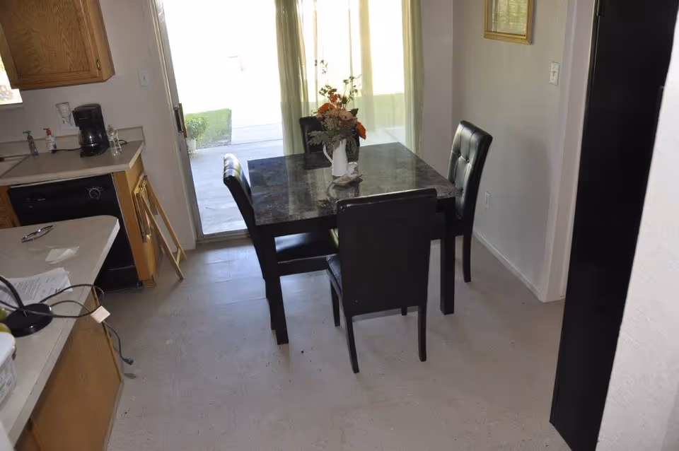 A small dining area with a square black table and four black chairs. A white vase with flowers is placed on the table. The dining area is adjacent to a kitchen with wooden cabinets, a coffee maker, and a dishwasher. A sliding glass door with sheer curtains leads to an outdoor space.