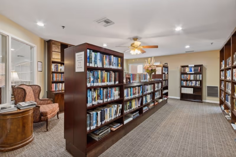 Interior view of a library room in a senior living facility with multiple wooden bookshelves filled with books. There is a comfortable upholstered armchair and a round wooden side table with a lamp and some magazines on it. The room has beige walls, carpeted floor, and a ceiling fan with lights.