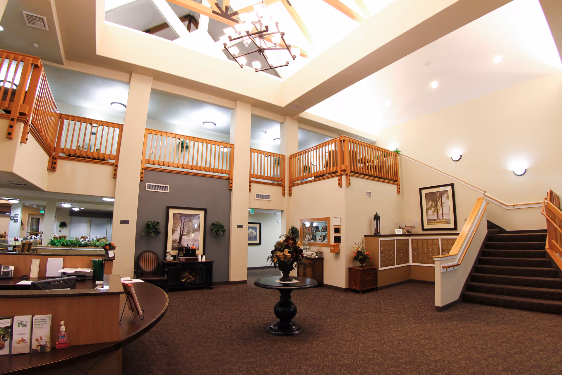 Spacious and well-lit senior living facility lobby with a high ceiling and wooden railings on the upper level. The area features a round table with a floral arrangement in the center, a staircase leading to the upper floor, a reception desk on the left, and framed artwork on the walls. The carpeted floor and warm lighting create a welcoming atmosphere.