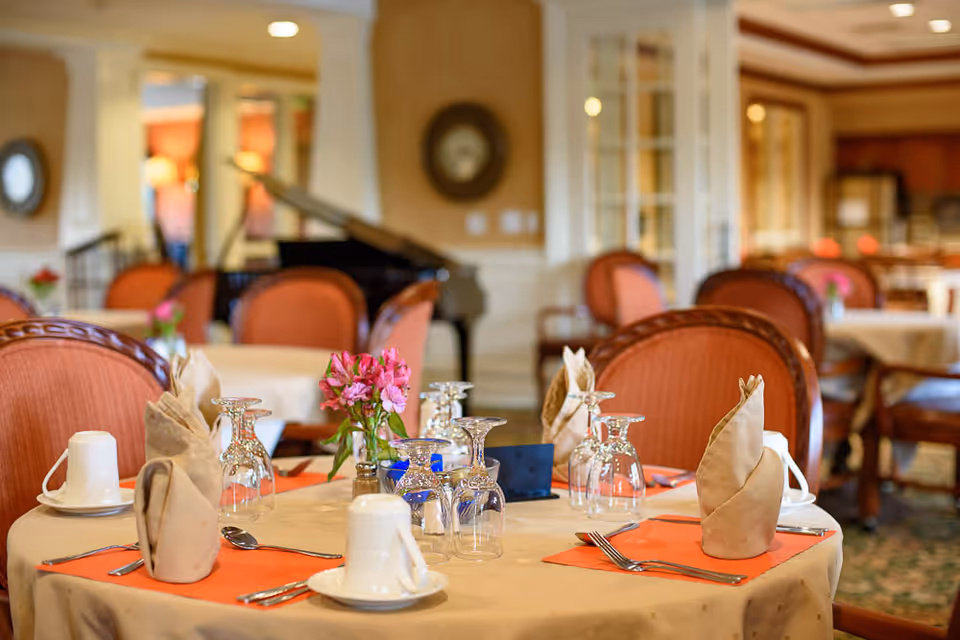 A dining room with round tables covered in beige tablecloths, each set with folded beige napkins, glassware, white cups, and silverware on orange placemats. Pink flowers in small vases decorate the tables. The room features upholstered chairs with wooden frames and a grand piano in the background.