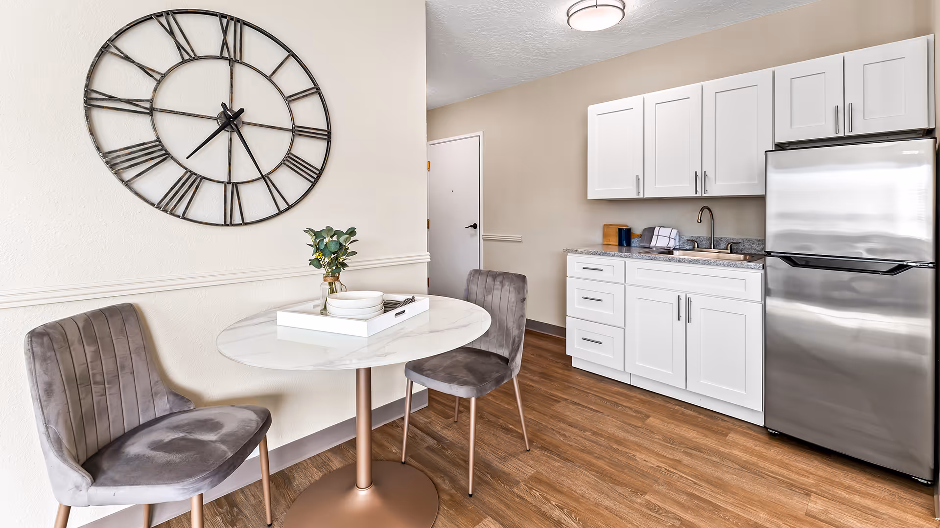 A small dining area and kitchenette in a senior living facility. The dining area features a round white table with a gold base, two gray upholstered chairs, and a decorative tray with dishes and a small plant. On the wall above the table is a large black metal clock with Roman numerals. The kitchenette has white cabinets, a granite countertop, a stainless steel refrigerator, and a sink. The floor is wood with a warm tone.