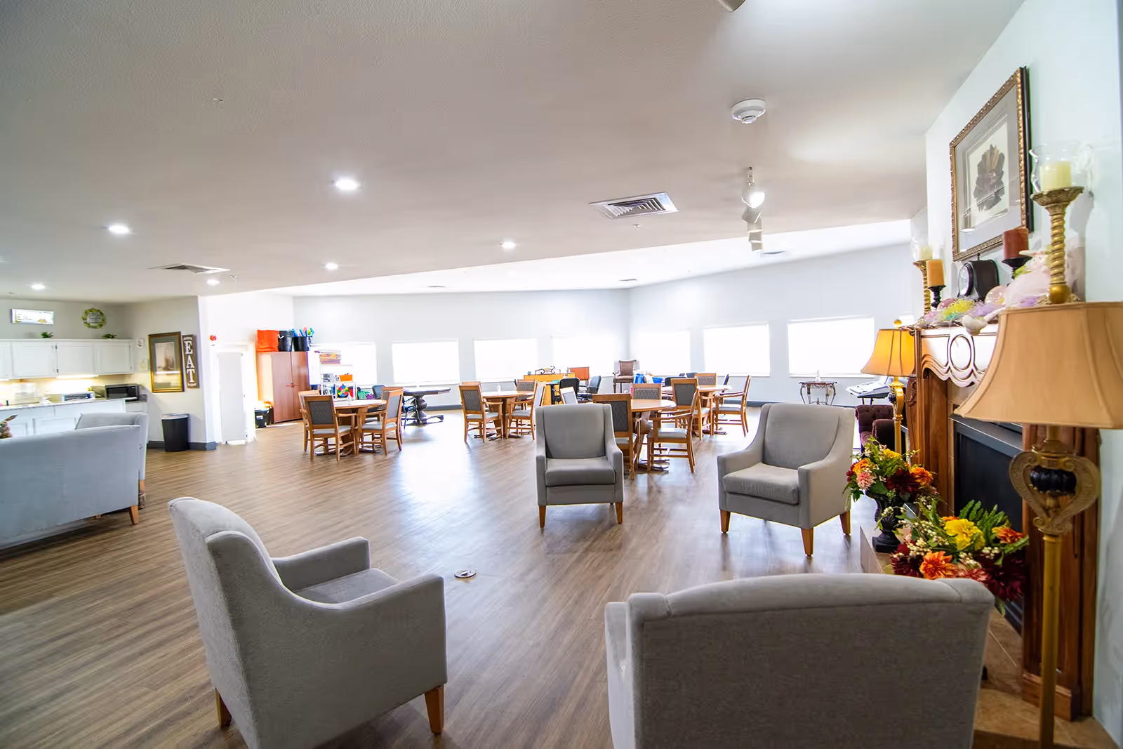 A spacious, well-lit common area with several gray armchairs arranged around a wooden fireplace decorated with candles and flowers. In the background, there are multiple wooden tables and chairs near large windows letting in natural light. The room has wood flooring and white walls, with a kitchen area visible to the left.