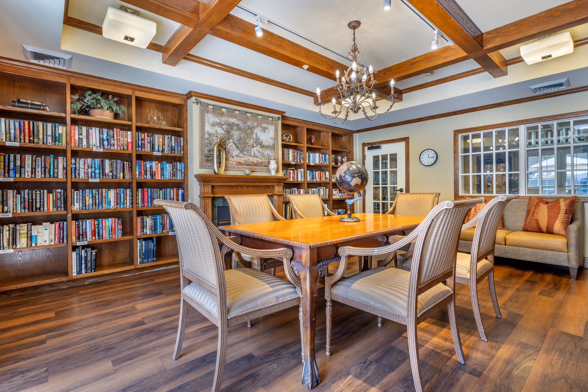 A cozy library or reading room with wooden bookshelves filled with books, a wooden table surrounded by six cushioned chairs, a globe on the table, a chandelier hanging from a ceiling with wooden beams, a fireplace with decorative items above it, a clock on the wall, and a sofa with pillows near a window.