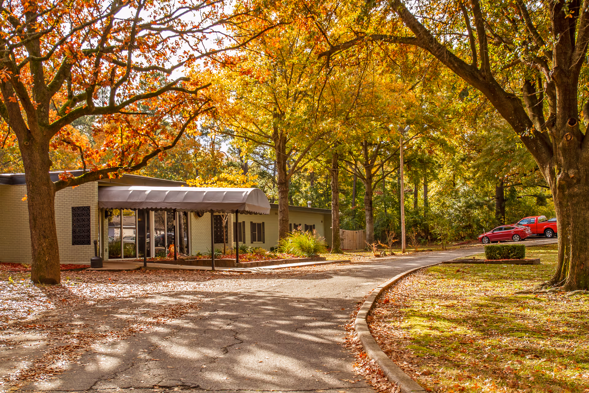 Exterior view of The Blossoms at Fort Smith Rehab & Nursing Center surrounded by trees with autumn foliage. The building has a covered entrance and a curved driveway leading to a parking area with a few cars.