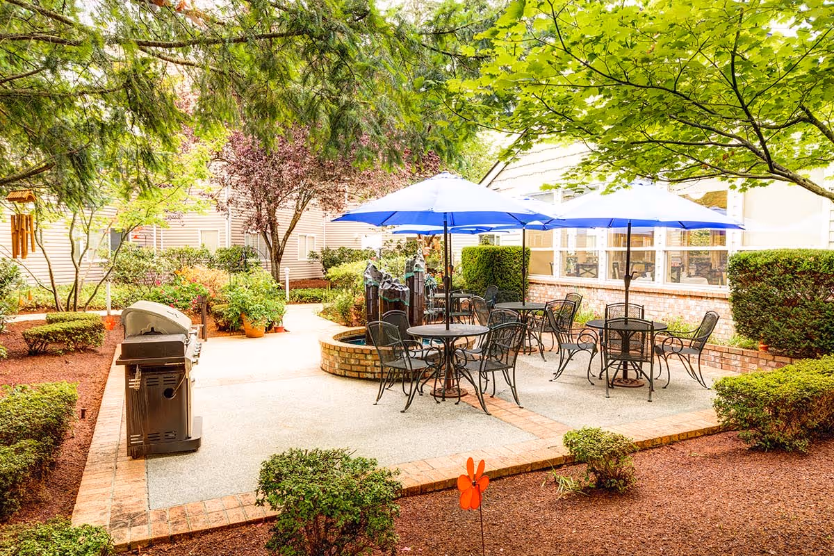 Outdoor patio area at The Gardens at Marysville with metal tables and chairs under blue umbrellas, surrounded by green trees, shrubs, and a brick-lined walkway. A barbecue grill is visible on the left side.
