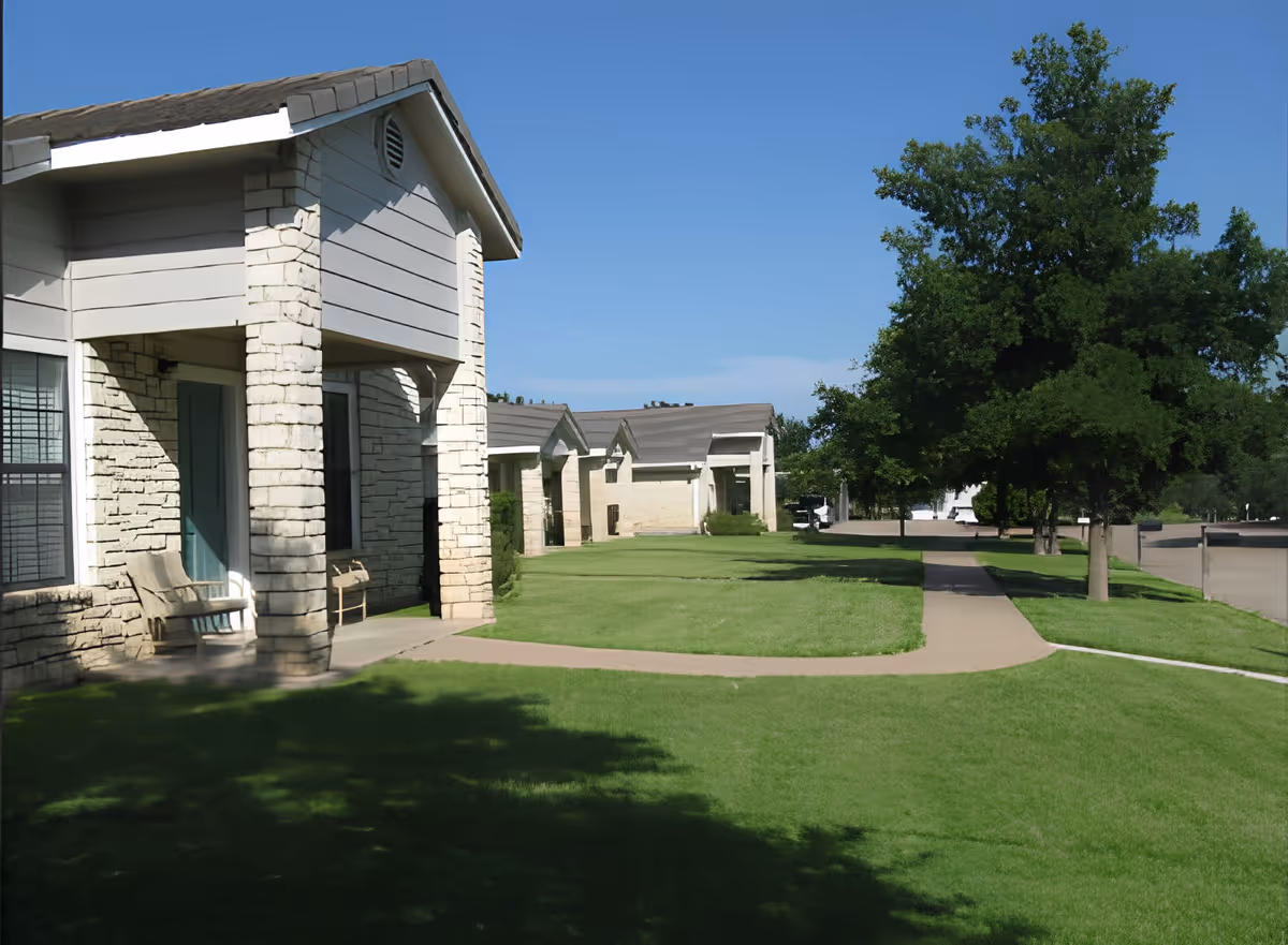 Exterior view of Marble Falls Assisted Living showing a row of single-story buildings with stone and siding facades, a well-maintained green lawn, a paved walkway, and several trees under a clear blue sky.