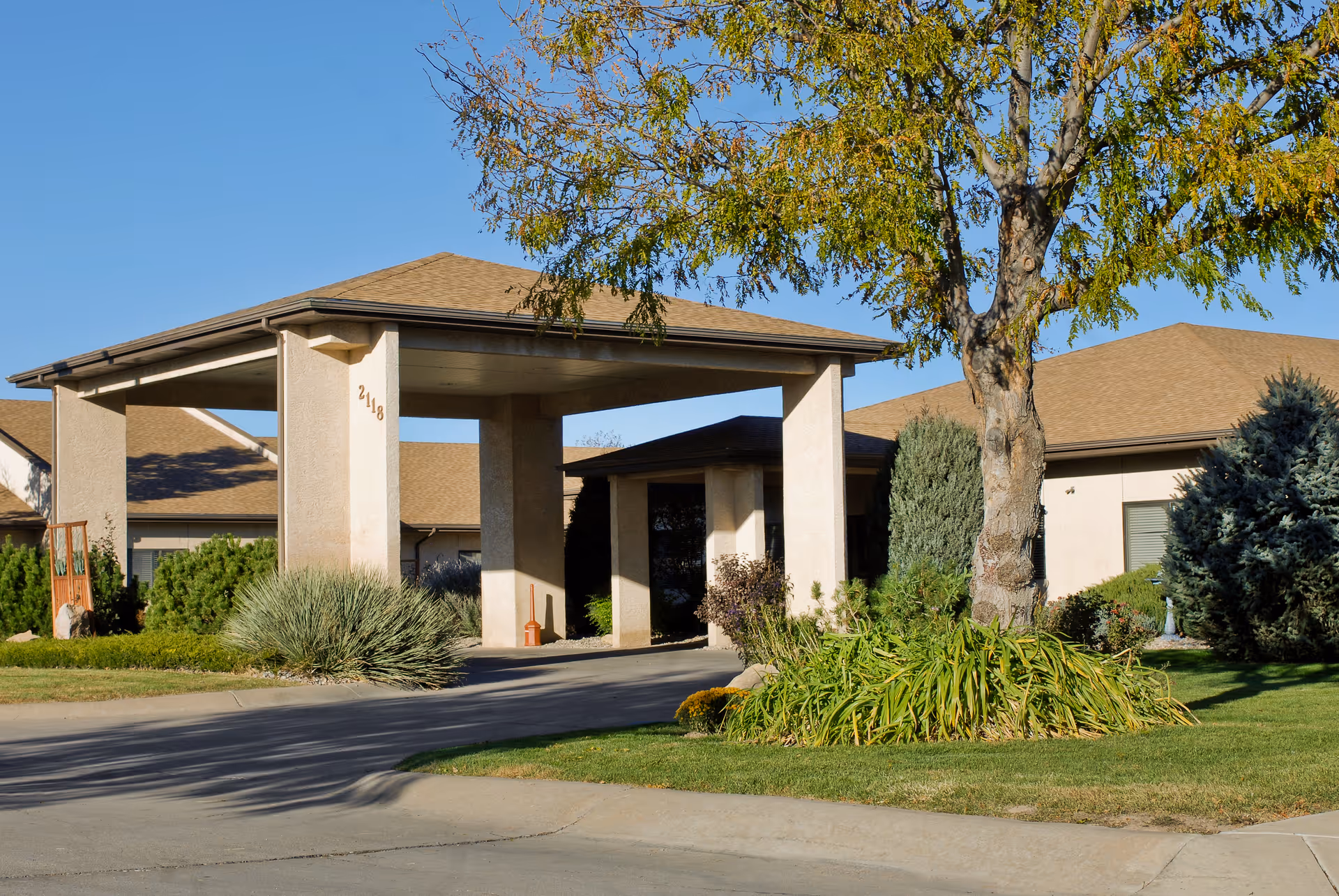 Exterior view of the Life Care Center of Pueblo showing the entrance with a covered driveway, surrounded by green shrubs, trees, and a well-maintained lawn under a clear blue sky.