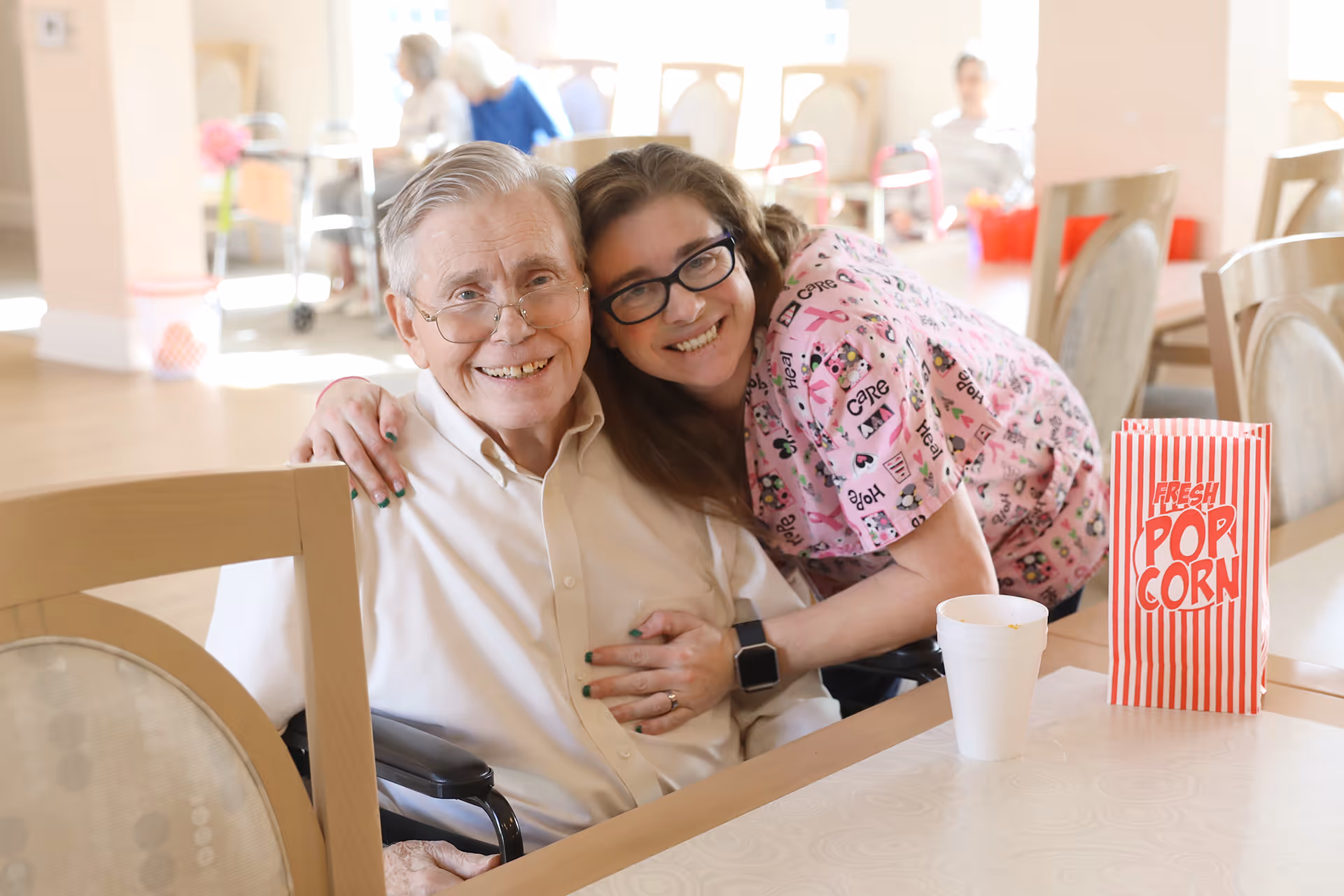 An elderly man in a wheelchair smiling as a caregiver hugs him at a dining table with a striped popcorn bag.