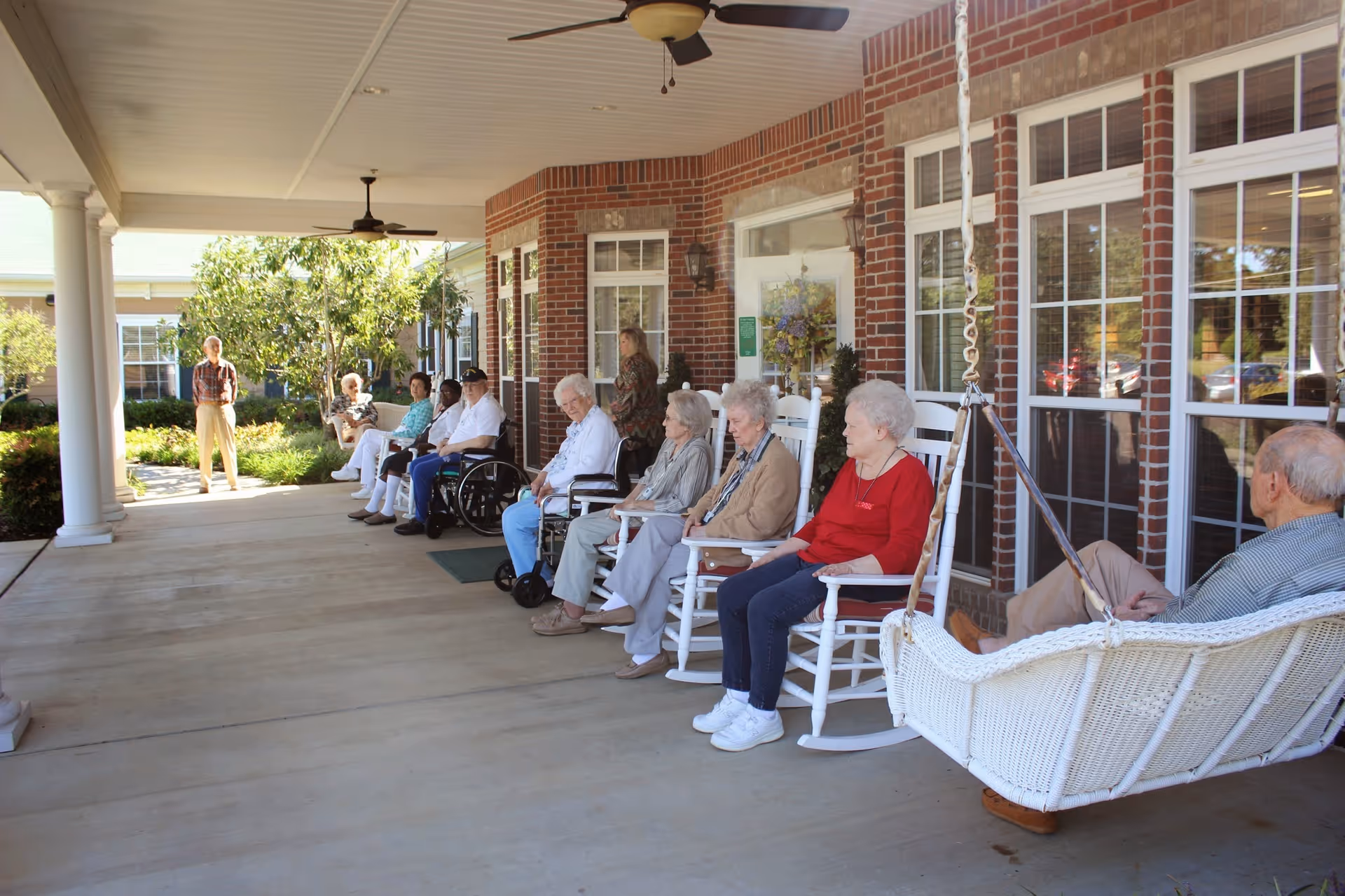 A group of elderly people sitting on rocking chairs and a swing on a covered porch outside a brick building, with a woman standing nearby and greenery in the background.
