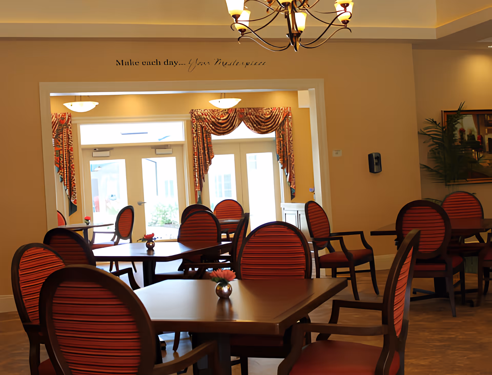 Interior view of a dining room with several wooden tables and red cushioned chairs. The room has beige walls, decorative curtains on the windows, and a chandelier hanging from the ceiling. A small flower vase is placed on each table. There is a wall-mounted hand sanitizer dispenser and a framed picture with a plant on the right side.