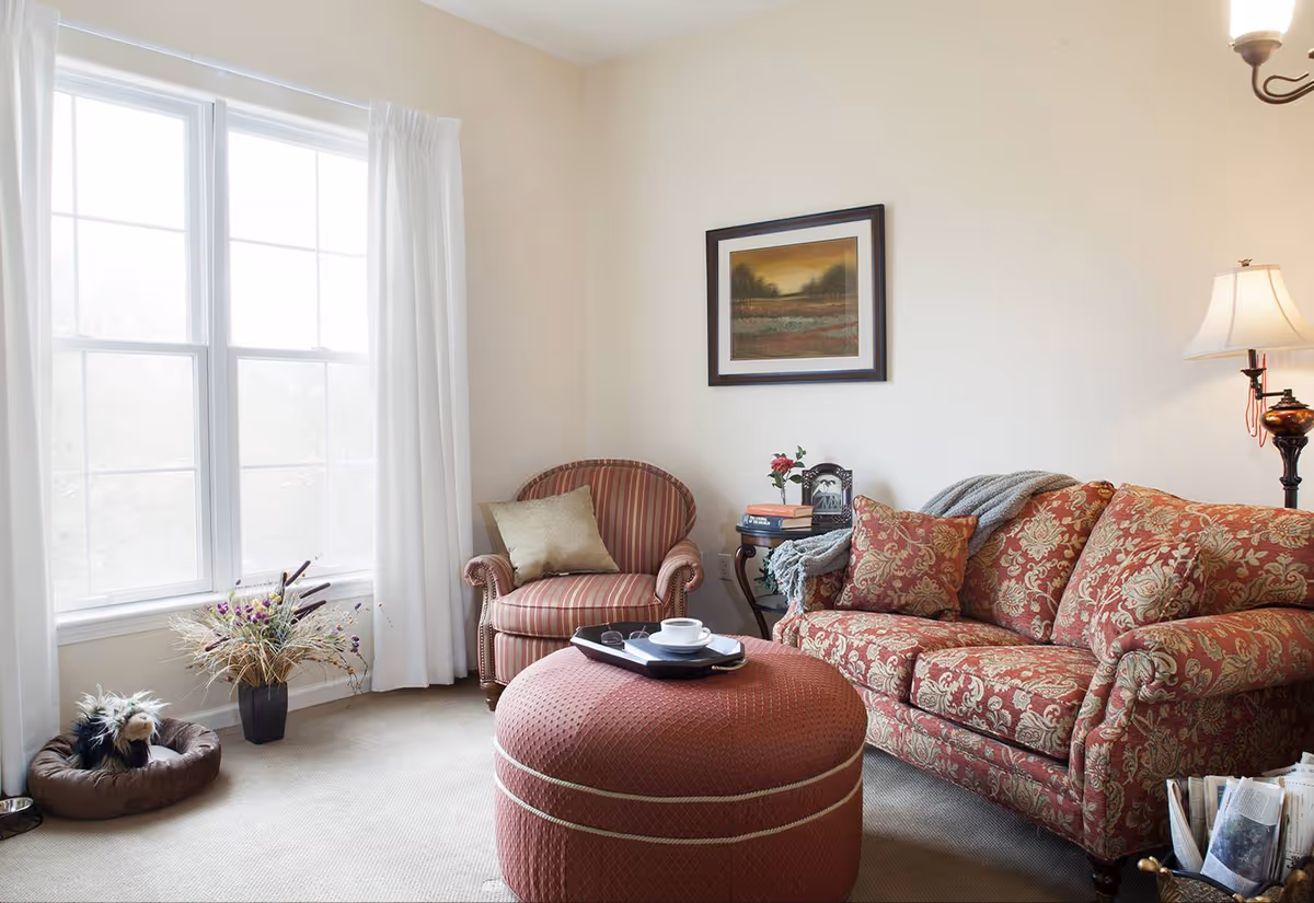 A cozy living room with a large window covered by white curtains, a patterned red sofa with matching cushions, a striped armchair with a beige pillow, and a round red ottoman with a tray holding a cup and saucer. A side table with books, a clock, and a small flower vase is next to the sofa. A floor lamp with a white shade stands behind the sofa. There is a dog bed with a small dog and a food bowl near the window, and a framed landscape painting hangs on the wall above the armchair.