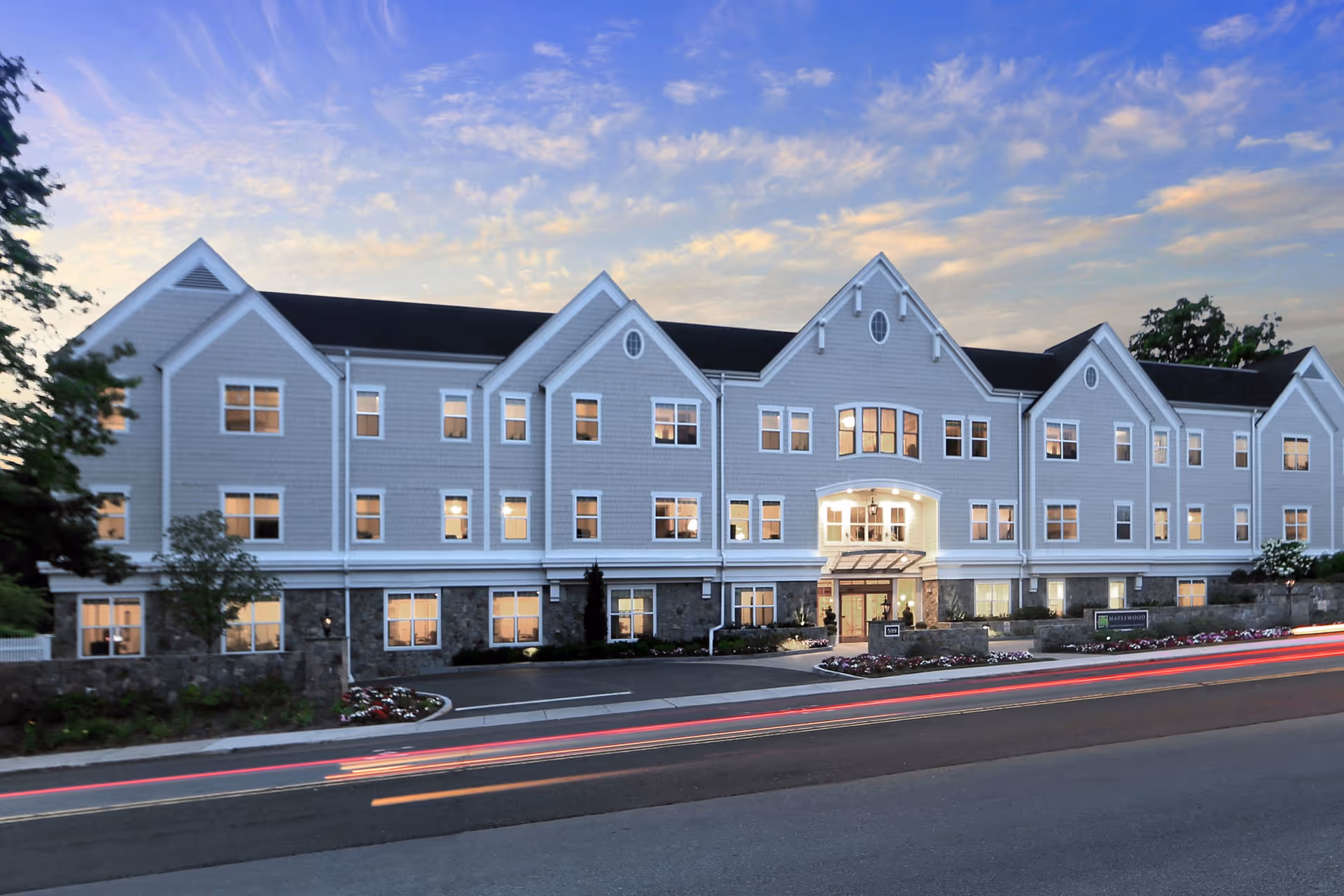 Exterior front view of a large, three-story senior living facility building named Maplewood at Darien during dusk, with lights glowing from the windows and a partly cloudy sky in the background.