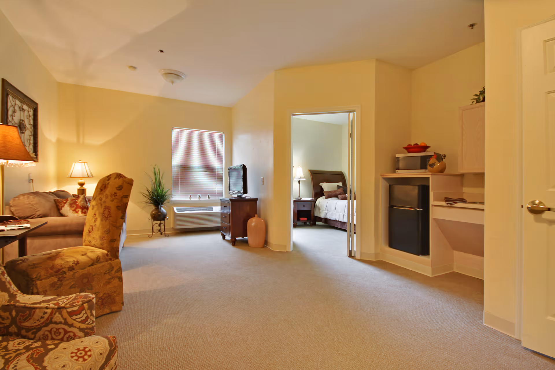 A cozy living area in a senior living facility with a sofa, two patterned armchairs, a side table with a lamp, a TV on a wooden stand, and a window with blinds. To the right, there is a small kitchenette area with a mini fridge, microwave, and countertop. An open doorway leads to a bedroom with a bed, nightstand, and lamp.