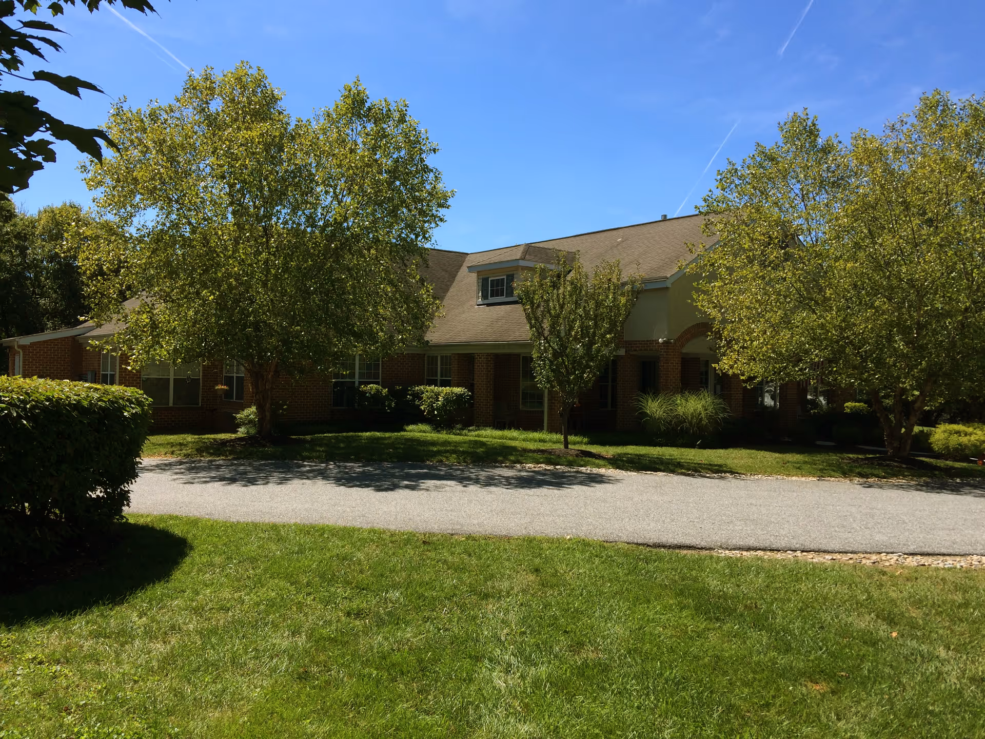 Front view of a one-story brick senior living building with trees, lawn, and a driveway under a clear blue sky.