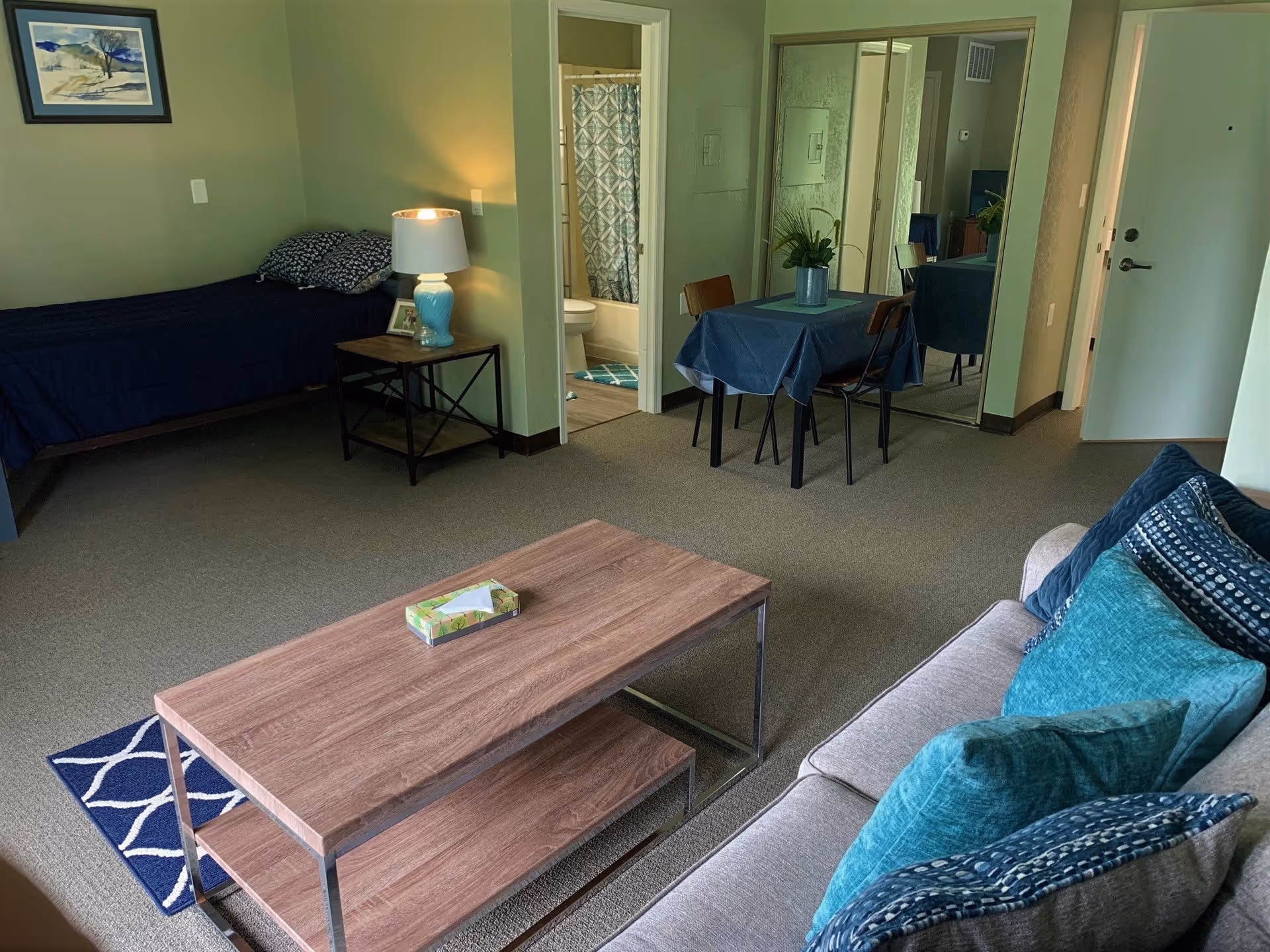 Interior view of a retirement community apartment showing a living area with a gray couch adorned with blue and teal pillows, a wooden coffee table with a tissue box, a small dining table with two chairs and a plant centerpiece, a bed with dark blue bedding and patterned pillows, a bedside table with a lamp, and a bathroom with a shower curtain visible through an open door.