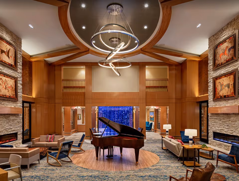 A spacious and elegant living room area with a grand piano in the center on a circular wooden platform. The room features modern lighting fixtures hanging from a high ceiling with wooden beams. There are multiple seating arrangements with sofas, armchairs, and side tables around the room. The walls are decorated with framed artwork and stone accents, and there are two modern fireplaces on either side. The overall ambiance is warm and inviting.